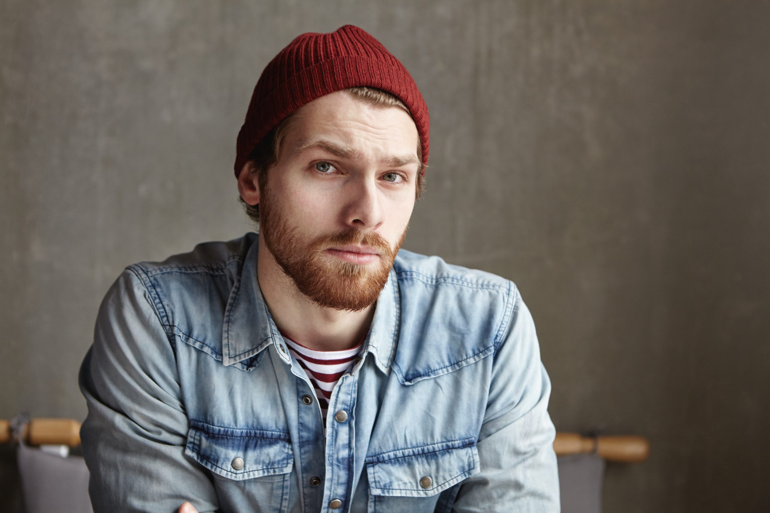 Headshot of macho hipster with stylish beard wearing hat and jeans shirt looking at camera with doubtful indecisive expression, feeling frustrated and puzzled about taking important decision